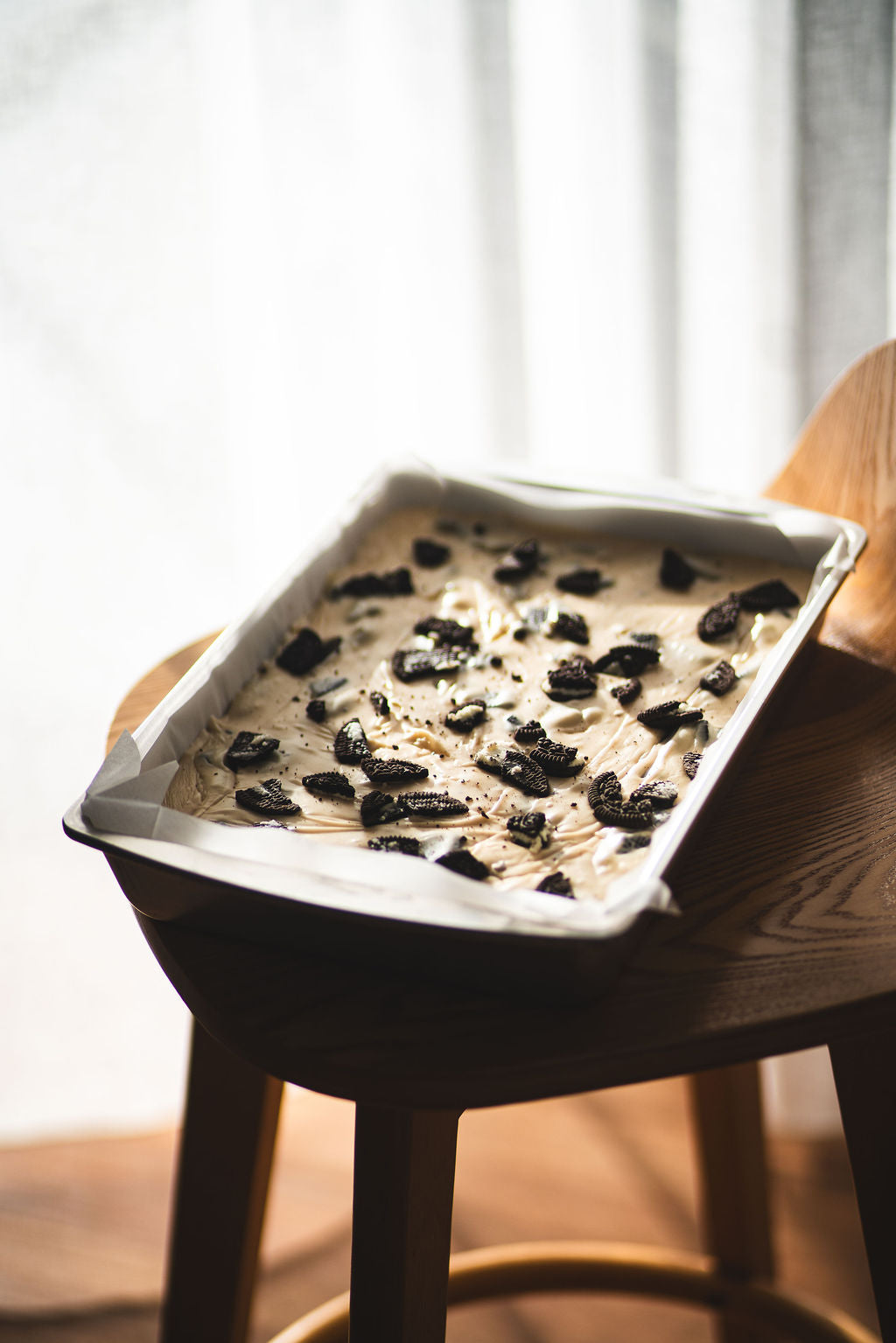 Tray of cookies and cream fudge topped with crushed chocolate biscuits, resting on a wooden chair in soft natural light.