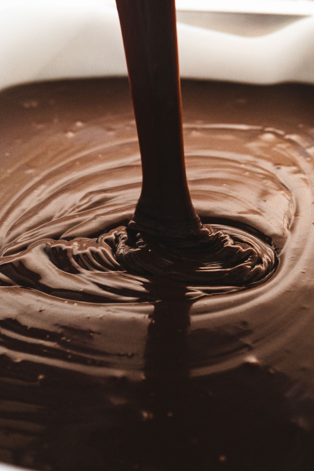 A close-up shot of rich, melted chocolate fudge being poured into a tray. The stream of glossy chocolate creates swirling ripples across the surface, highlighting its smooth, velvety texture. The warm lighting emphasizes the depth and indulgence of the fudge.