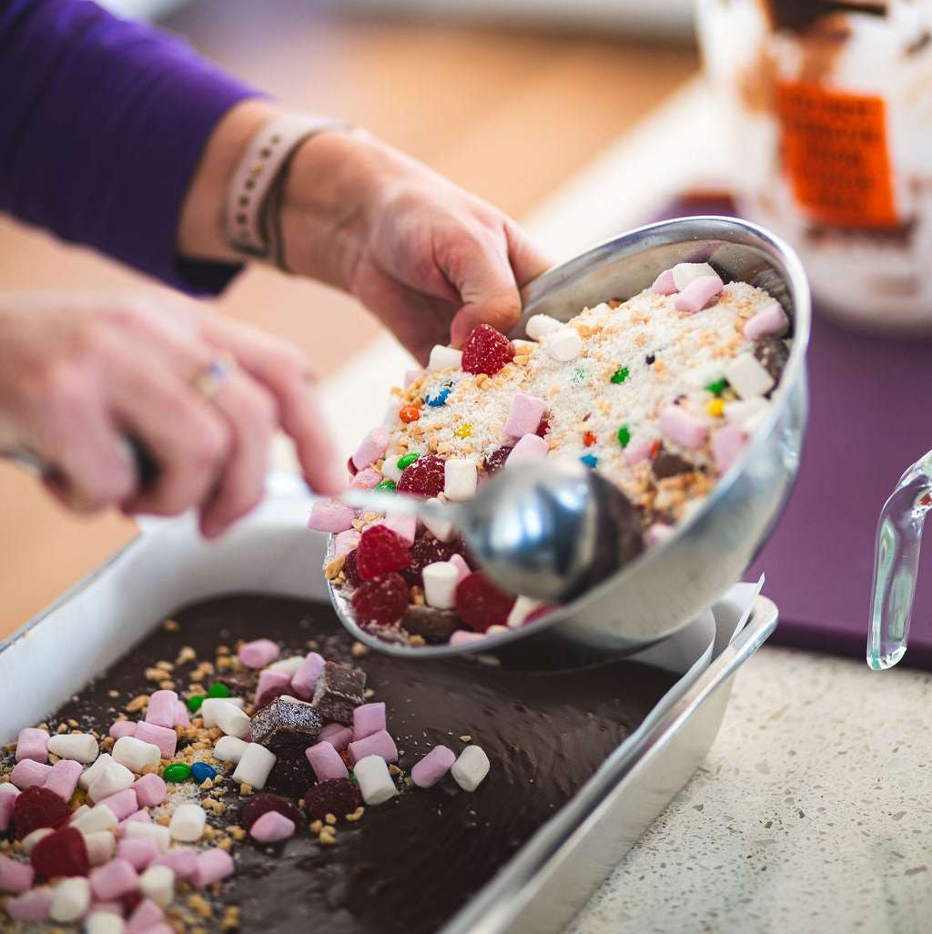 Hands topping a tray of chocolate fudge with marshmallows, raspberries, and colorful candy mix.