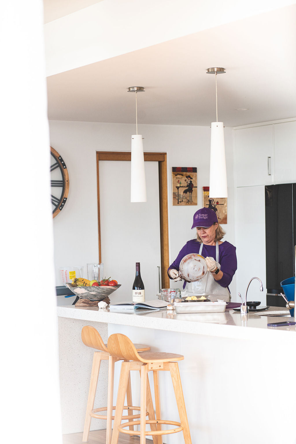 Woman in a purple shirt and cap pouring chocolate mixture into a tray in a bright kitchen.
