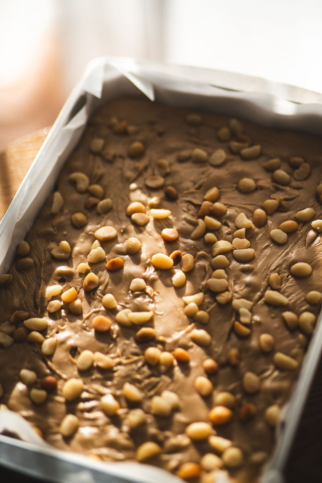 Tray of creamy caramel fudge topped with roasted macadamia nuts in warm natural light.