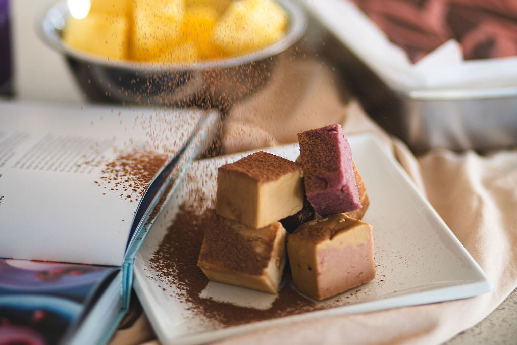 Close-up of assorted fudge cubes on a white plate as cocoa powder is sprinkled over them.