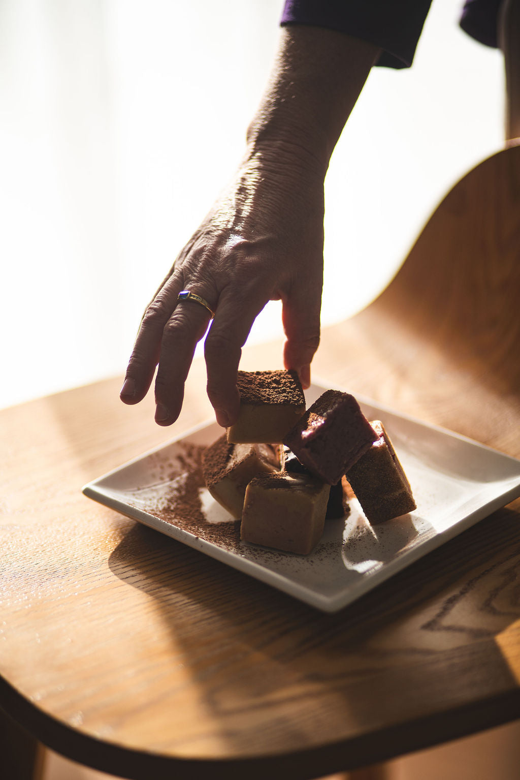 Hand reaching to pick up assorted fudge cubes stacked on a white plate dusted with cocoa, sitting on a wooden chair in soft natural light.