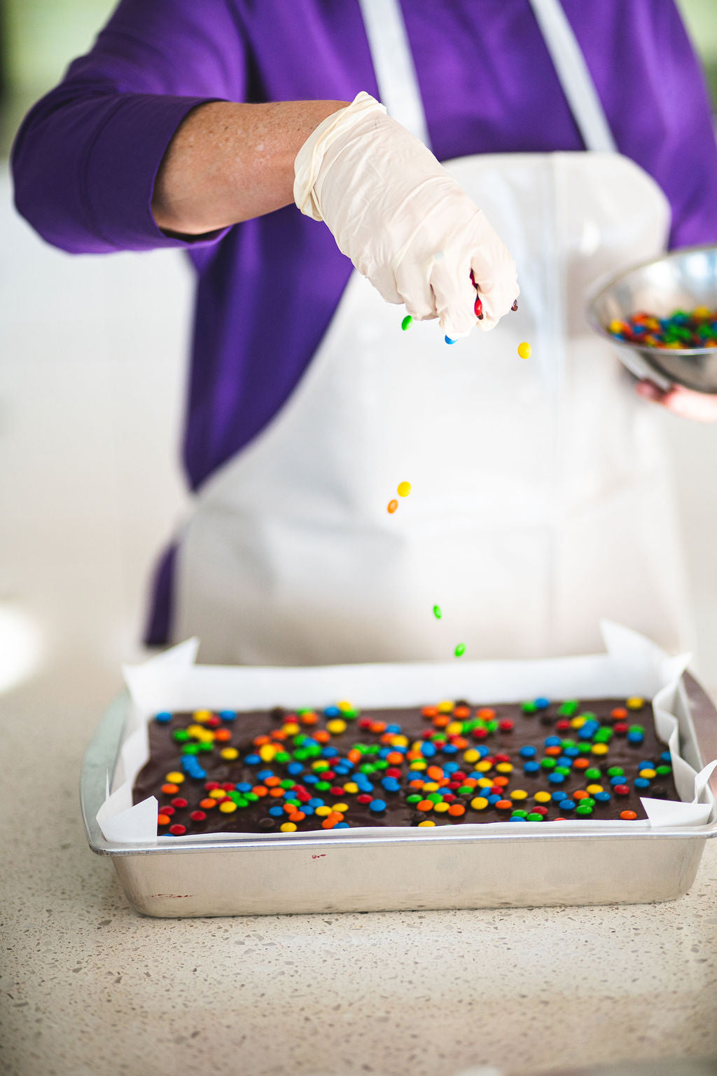 Person sprinkling colorful candy pieces over a tray of chocolate fudge.