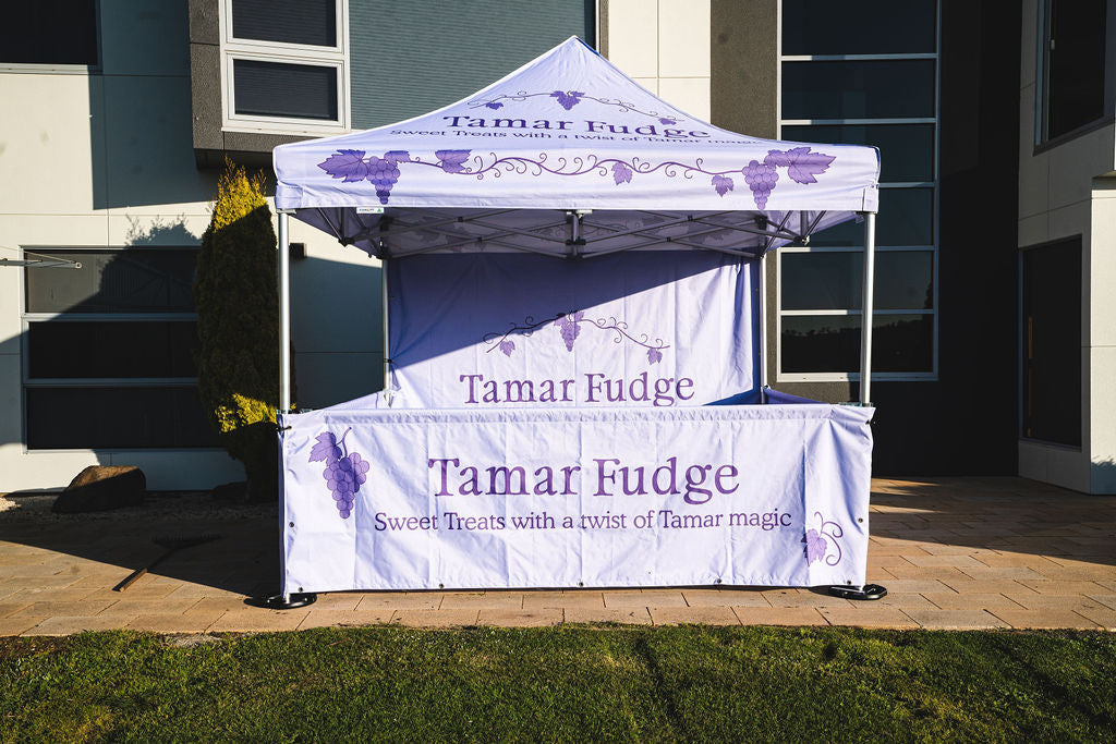 Lavender-purple Tamar Fudge market stall tent set up outdoors, branded with grape illustrations and the slogan ‘Sweet Treats with a twist of Tamar magic.’